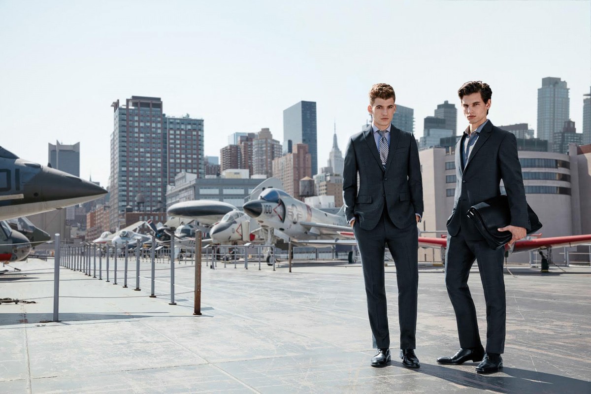 Two adult men in suits stand on an aircraft carrier deck in front of vintage planes with a cityscape in the background on a sunny day.