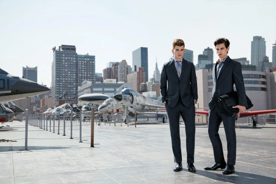 Two adult men in suits stand on an aircraft carrier deck in front of vintage planes with a cityscape in the background on a sunny day.