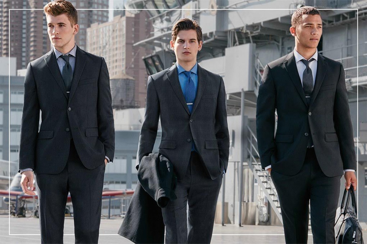 Three men stand outdoors in dark suits with ties and dress shirts against a modern cityscape backdrop, exuding a formal style.
