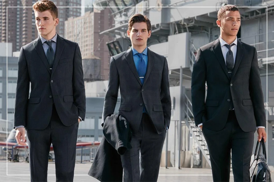 Three men stand outdoors in dark suits with ties and dress shirts against a modern cityscape backdrop, exuding a formal style.
