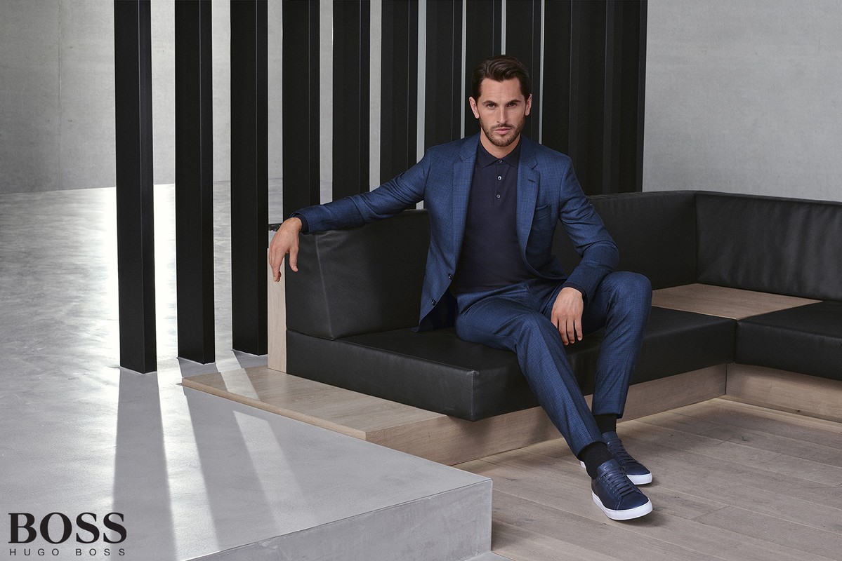 A man in a blue suit sits on a black couch in a studio. The man wears a blue polo shirt and blue sneakers. The setting is minimalist and modern.
