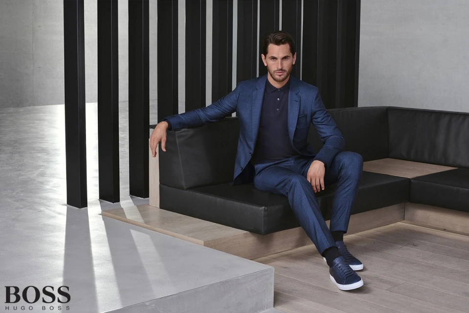 A man in a blue suit sits on a black couch in a studio. The man wears a blue polo shirt and blue sneakers. The setting is minimalist and modern.