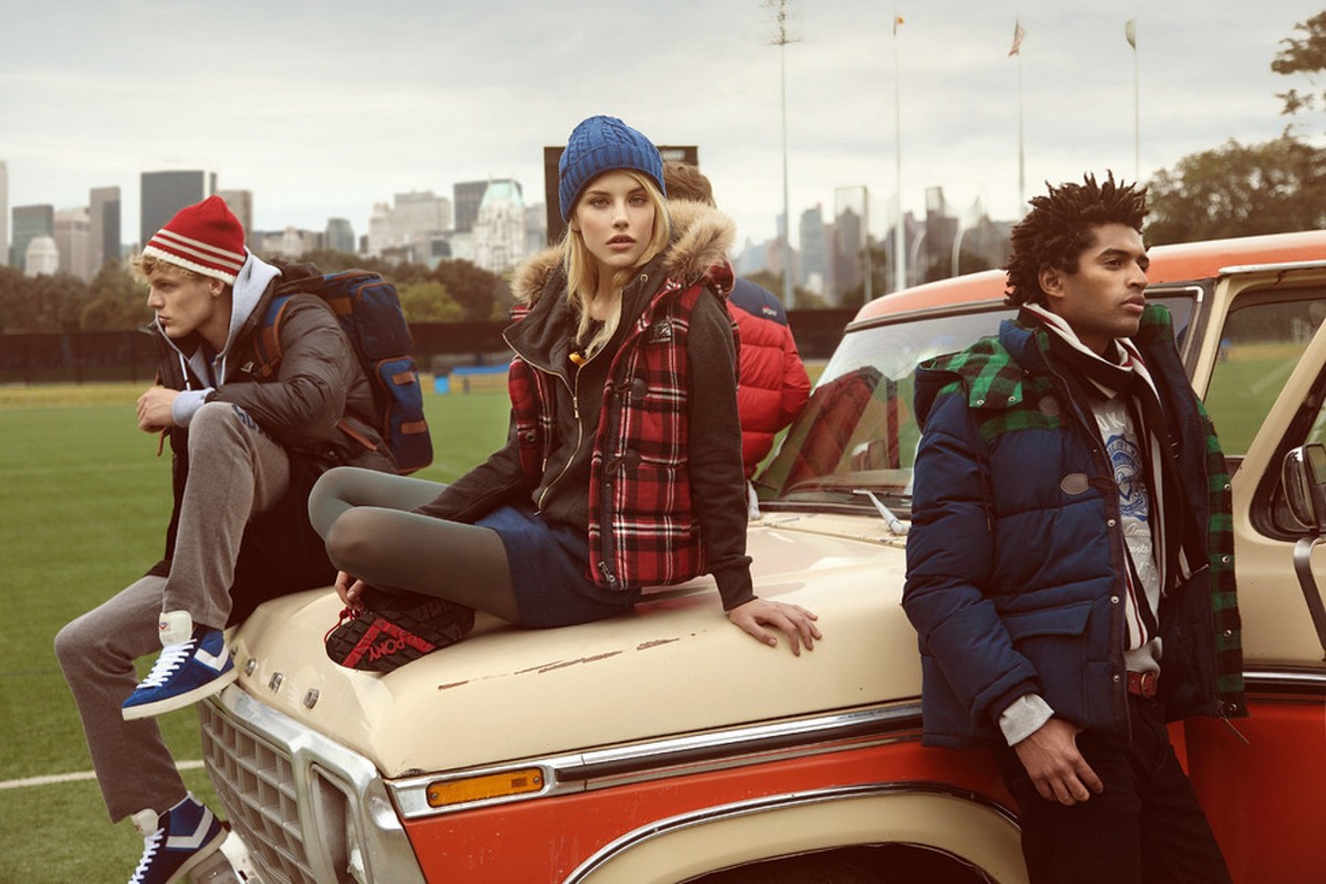 Three fashion models pose on a vintage truck parked on a grassy field with an urban skyline in the background, showcasing outerwear.