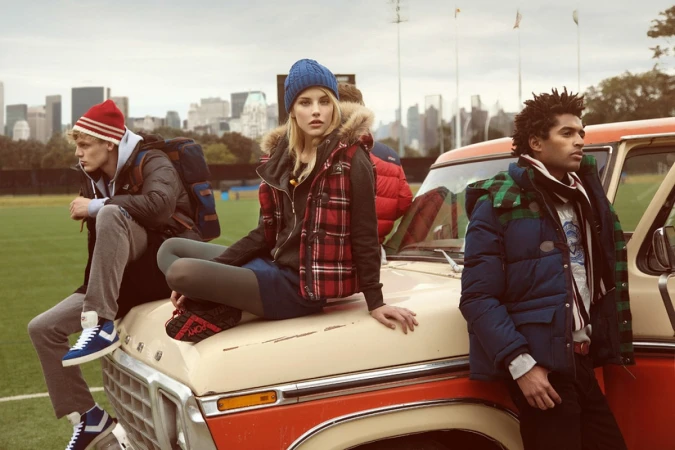 Three fashion models pose on a vintage truck parked on a grassy field with an urban skyline in the background, showcasing outerwear.