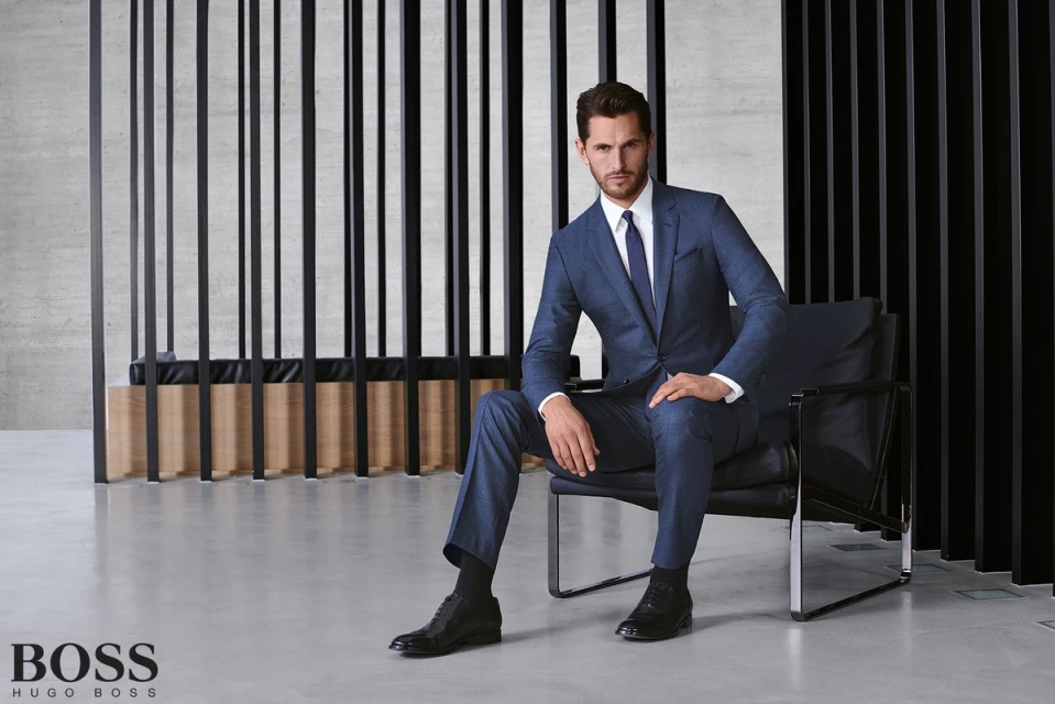 An adult male fashion model in a blue suit and tie, sitting in a black chair in a studio with black and beige decorative panels.