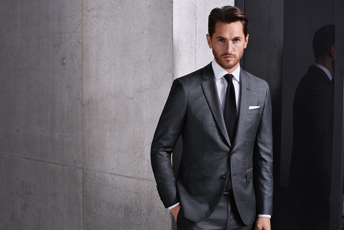 A stylish adult male in a grey suit and black tie poses indoors against a textured grey wall and sleek black reflective background.