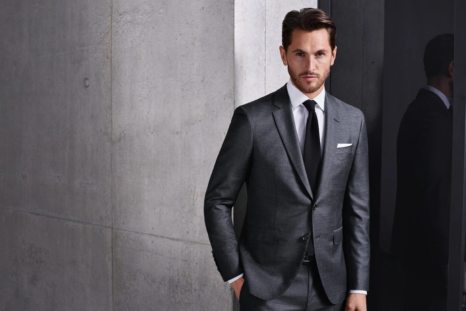 A stylish adult male in a grey suit and black tie poses indoors against a textured grey wall and sleek black reflective background.