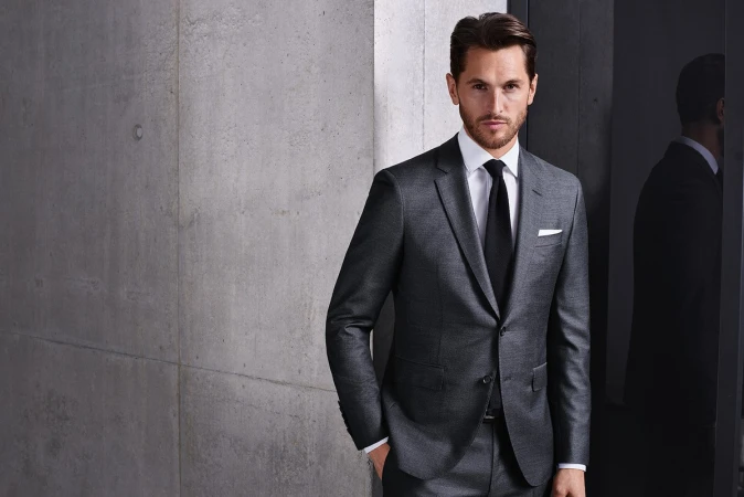 A stylish adult male in a grey suit and black tie poses indoors against a textured grey wall and sleek black reflective background.