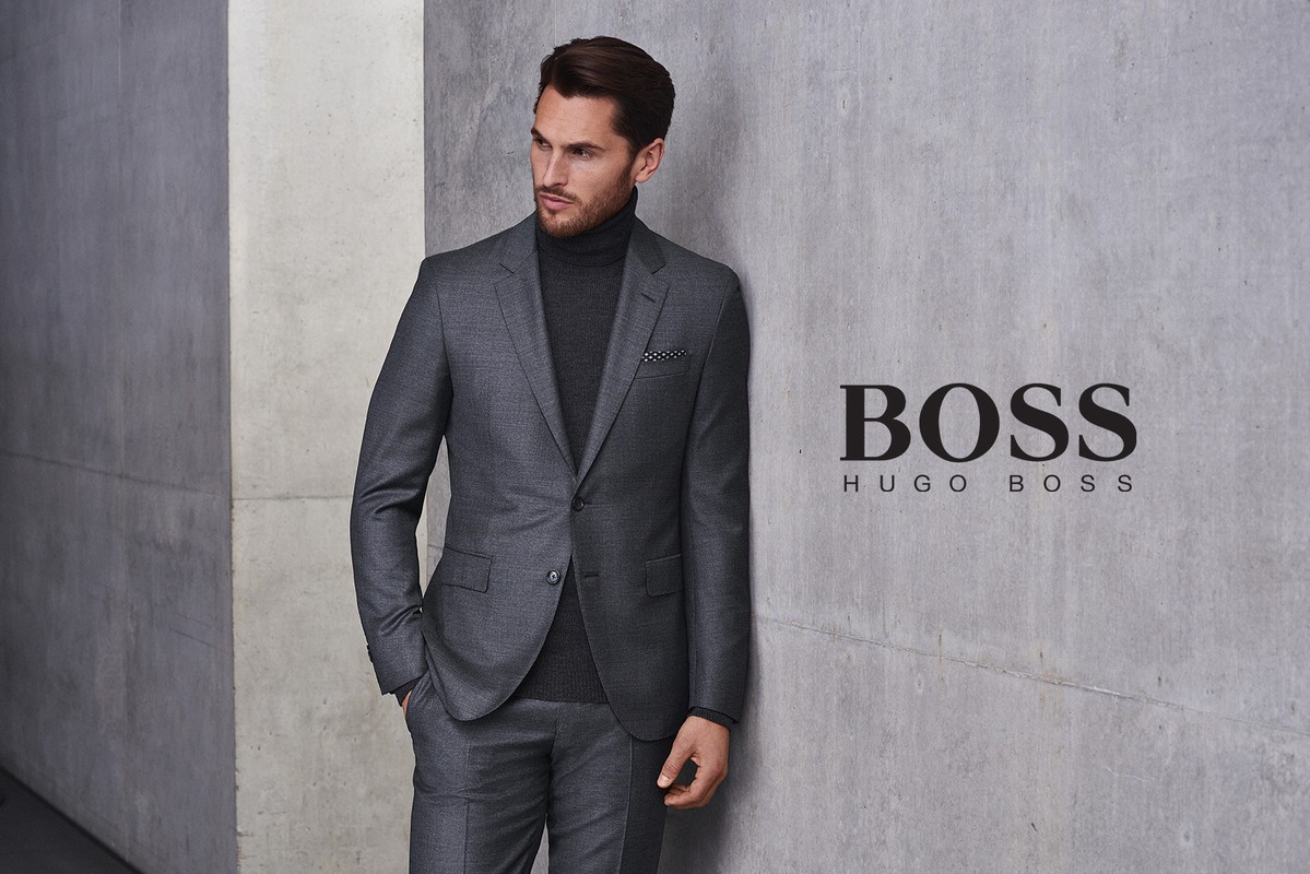 Adult male in a grey suit with a turtleneck poses against a grey wall with the Hugo Boss logo. Fashion advertisement.
