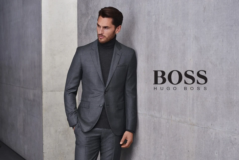 Adult male in a grey suit with a turtleneck poses against a grey wall with the Hugo Boss logo. Fashion advertisement.