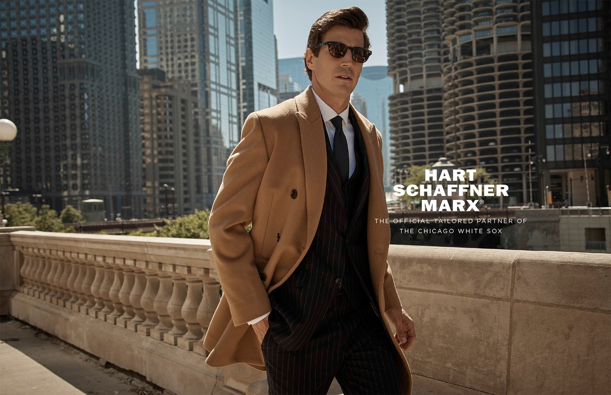 Man in tan overcoat, black suit, and sunglasses poses in urban setting with Chicago buildings in background on sunny day.