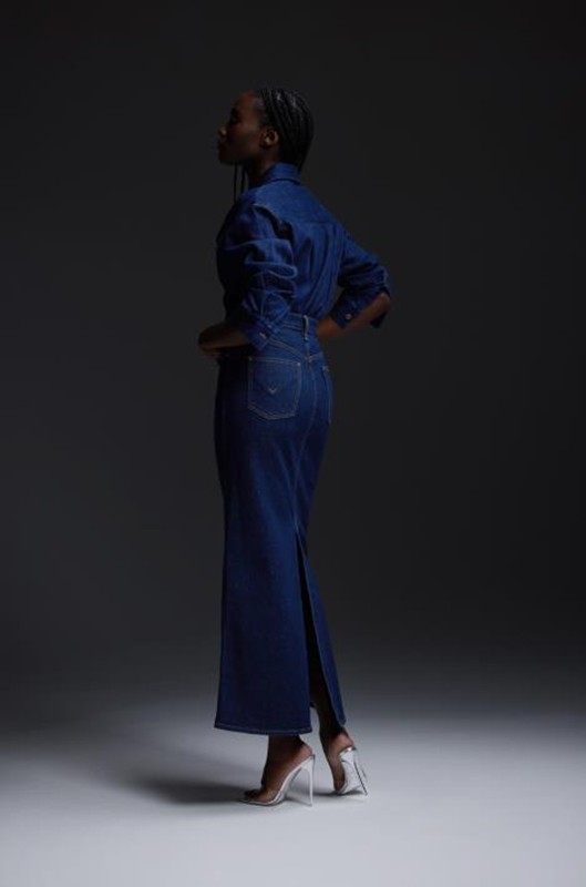 Woman in a denim dress and clear heels standing in a studio setting against a dark gray background, showcasing fashion and style.