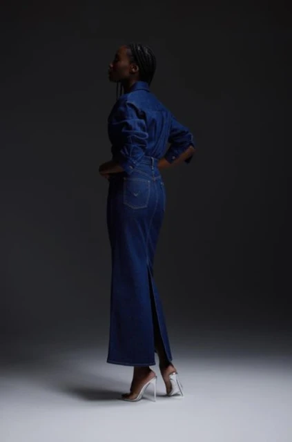 Woman in a denim dress and clear heels standing in a studio setting against a dark gray background, showcasing fashion and style.