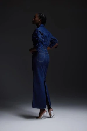 Woman in a denim dress and clear heels standing in a studio setting against a dark gray background, showcasing fashion and style.