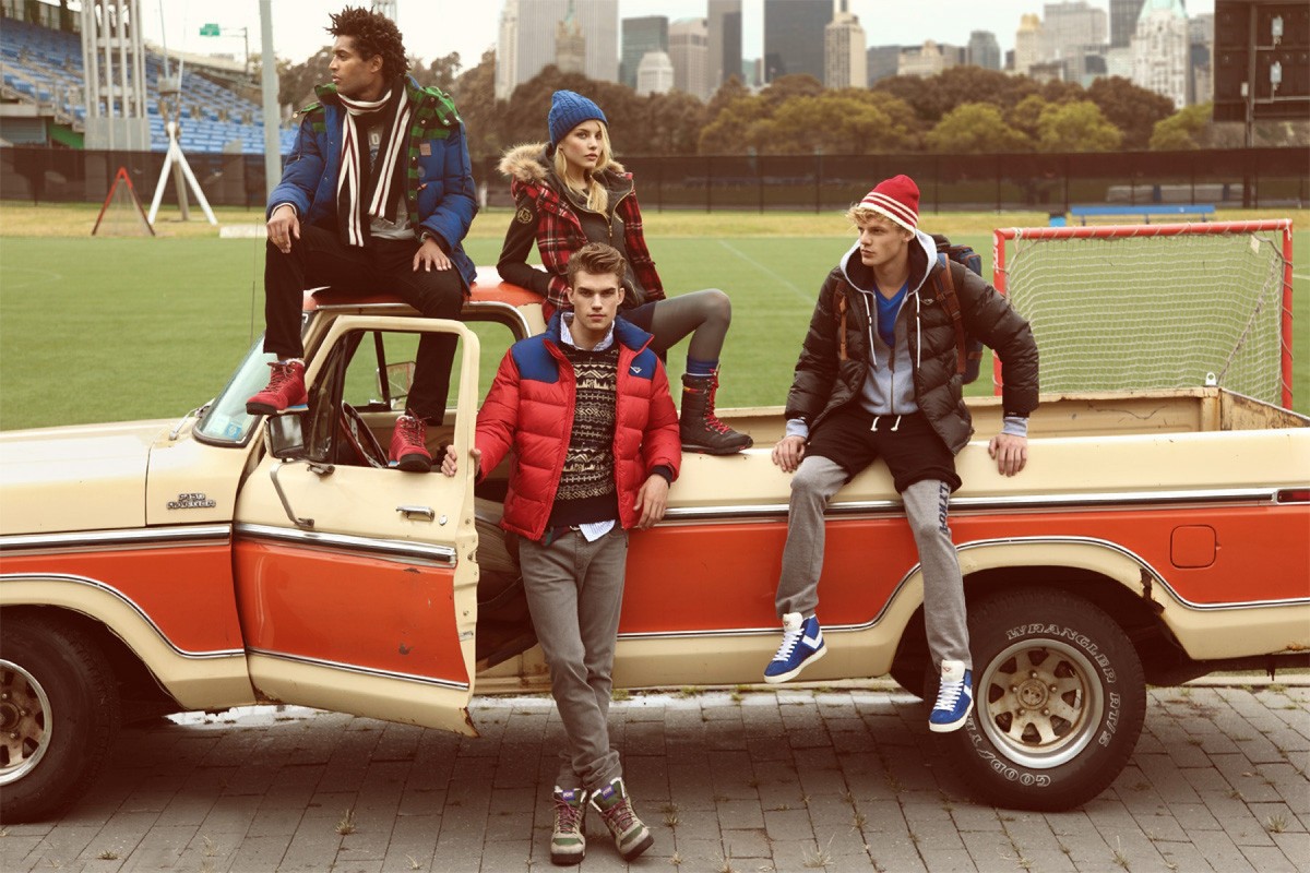 Four young adults pose on and around an orange and cream pickup truck, with a city skyline and sports field in the background.