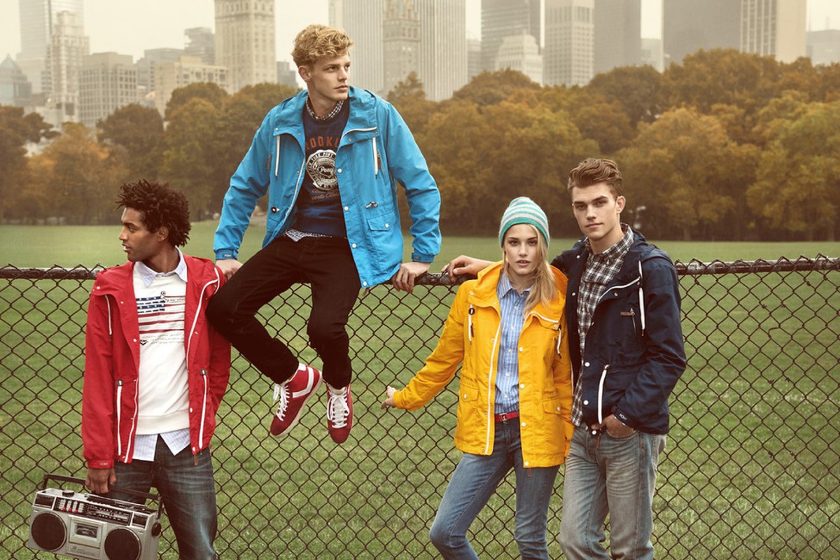 Four young adult models pose outdoors with the urban skyline behind them. They wear brightly colored jackets and casual clothing in an editorial style.