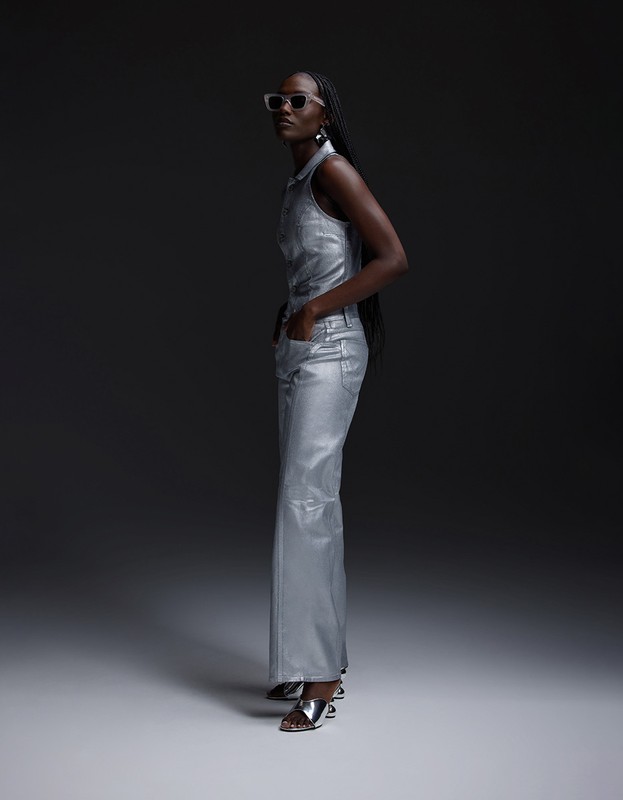 An adult female poses in a silver, sleeveless, collared shirt and pants with silver heels against a dark grey backdrop in a studio setting.