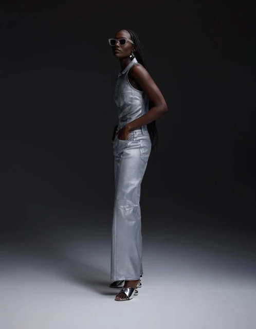 An adult female poses in a silver, sleeveless, collared shirt and pants with silver heels against a dark grey backdrop in a studio setting.