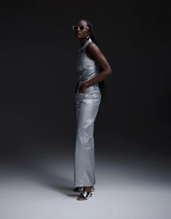An adult female poses in a silver, sleeveless, collared shirt and pants with silver heels against a dark grey backdrop in a studio setting.