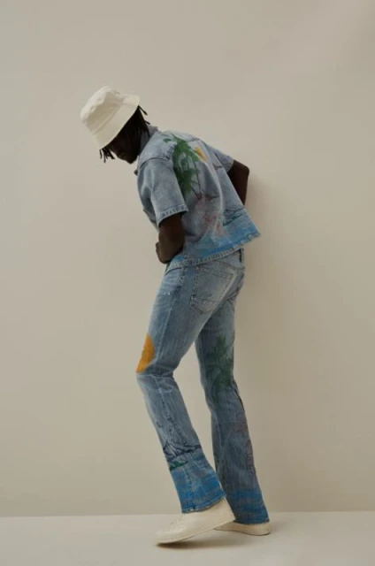 A dark-skinned adult male wearing a blue denim outfit with island motifs, posing indoors against an off-white wall. He's wearing a white bucket hat.