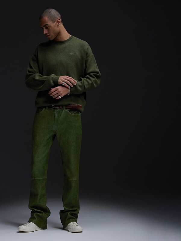 Man poses indoors in olive green sweater and pants with neutral sneakers in a fashion editorial studio shot against a dark background.