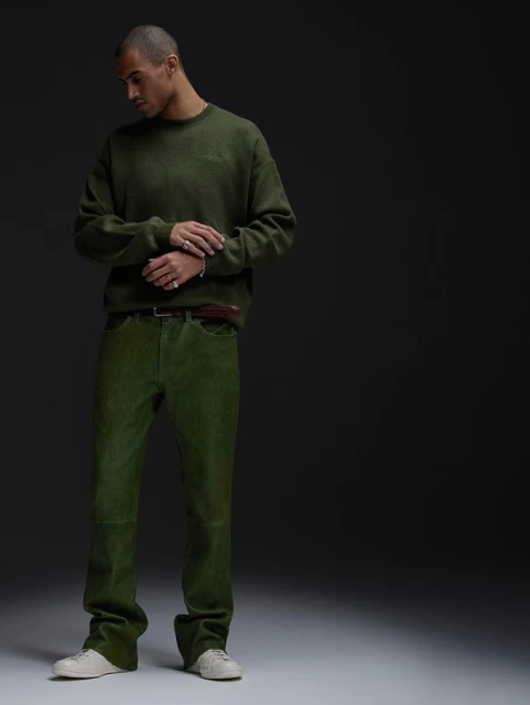 Man poses indoors in olive green sweater and pants with neutral sneakers in a fashion editorial studio shot against a dark background.