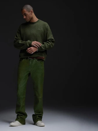 Man poses indoors in olive green sweater and pants with neutral sneakers in a fashion editorial studio shot against a dark background.