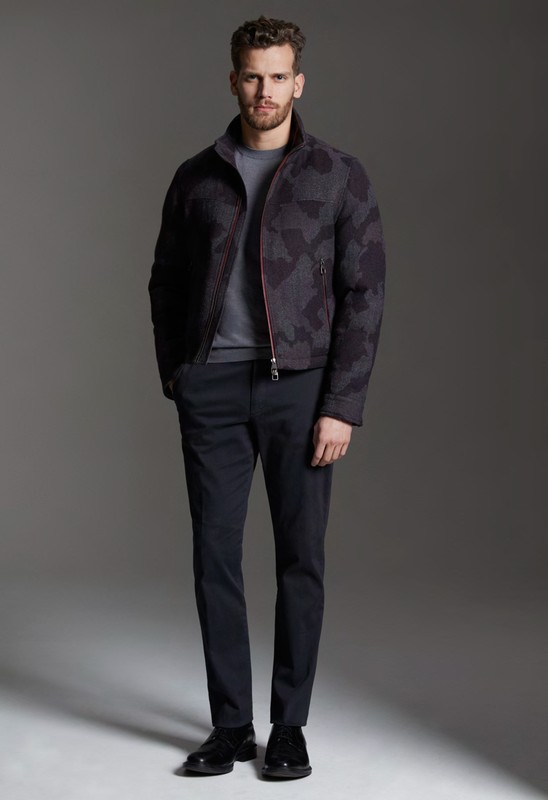 Full-length portrait of a male model wearing a dark patterned jacket, grey t-shirt, and trousers against a grey backdrop.