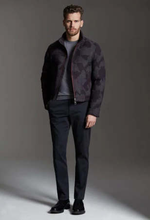 Full-length portrait of a male model wearing a dark patterned jacket, grey t-shirt, and trousers against a grey backdrop.