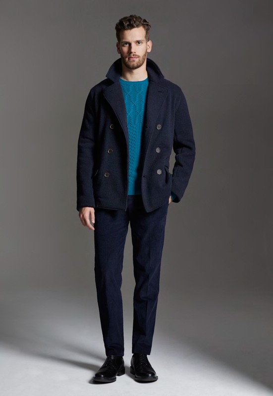 Full-length shot of an adult male fashion model wearing a blue double-breasted jacket, turquoise sweater, and blue pants against a grey backdrop.