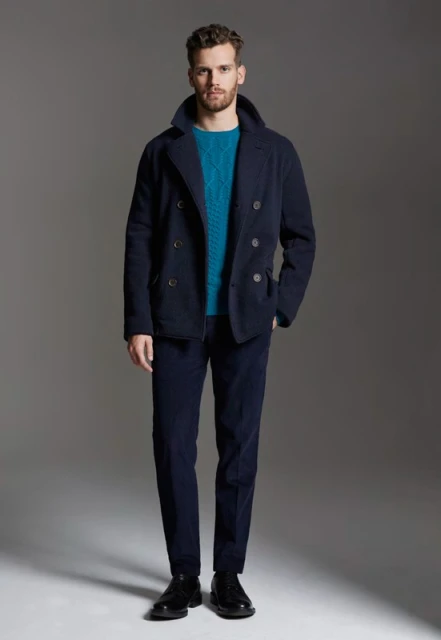 Full-length shot of an adult male fashion model wearing a blue double-breasted jacket, turquoise sweater, and blue pants against a grey backdrop.