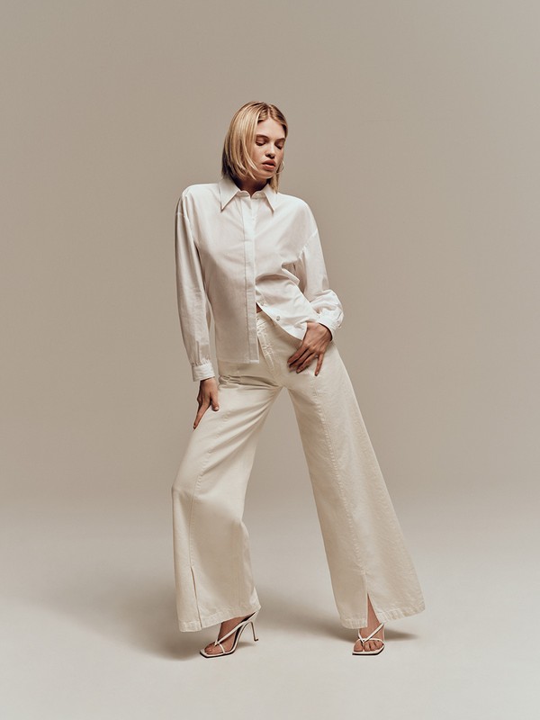 Blonde woman poses in white long-sleeved collared shirt and loose white pants with heels on a neutral-color indoor background.