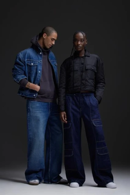 Two models, an adult male and female, stand indoors in an apparel fashion shot wearing denim jackets and wide-leg jeans on a gray background.