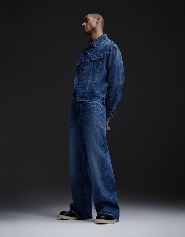 Man in denim jacket and wide-leg jeans poses in a studio with a gray background. Fashion-forward look with a modern, relaxed silhouette.