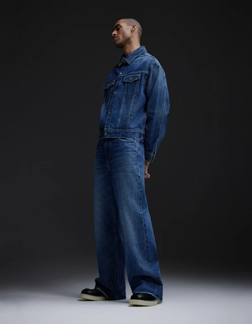Man in denim jacket and wide-leg jeans poses in a studio with a gray background. Fashion-forward look with a modern, relaxed silhouette.