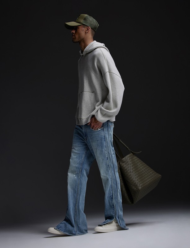 Man wearing a grey hoodie, light blue jeans, and a cap while holding a dark green tote bag in a studio