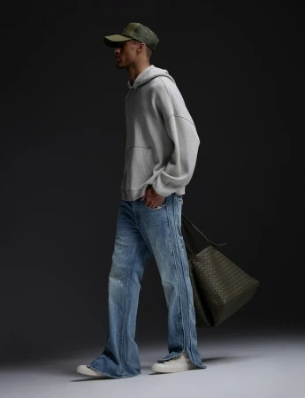 Man wearing a grey hoodie, light blue jeans, and a cap while holding a dark green tote bag in a studio