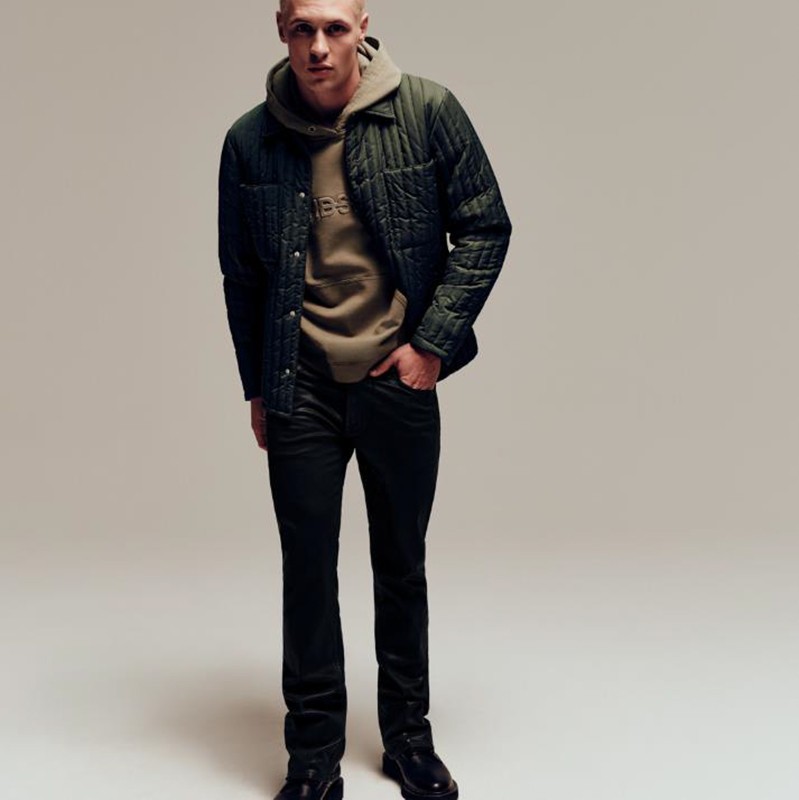 A slender adult male poses in dark jeans, black boots, and a green quilted jacket layered over a tan hooded sweatshirt in a studio shot.