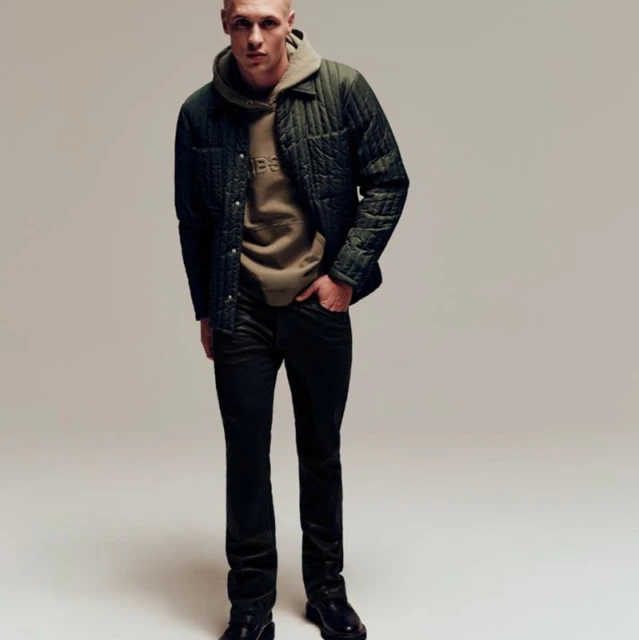 A slender adult male poses in dark jeans, black boots, and a green quilted jacket layered over a tan hooded sweatshirt in a studio shot.