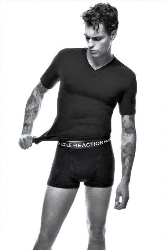 Black and white portrait of a young man wearing a black t-shirt and boxer briefs against a white background.