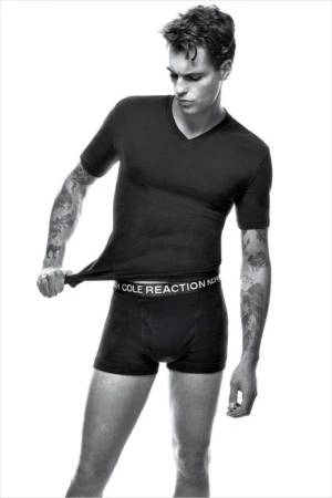 Black and white portrait of a young man wearing a black t-shirt and boxer briefs against a white background.