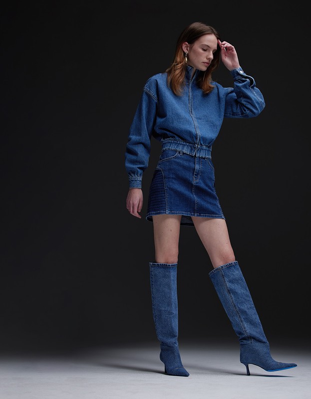 Model in full denim outfit including zip jacket, miniskirt, and knee-high boots on a grey backdrop