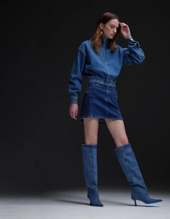 Model in full denim outfit including zip jacket, miniskirt, and knee-high boots on a grey backdrop