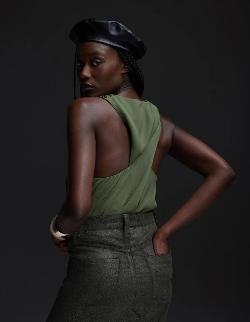 An African-American woman with an olive-green top and black beret looks back over her shoulder with a neutral expression in studio lighting.