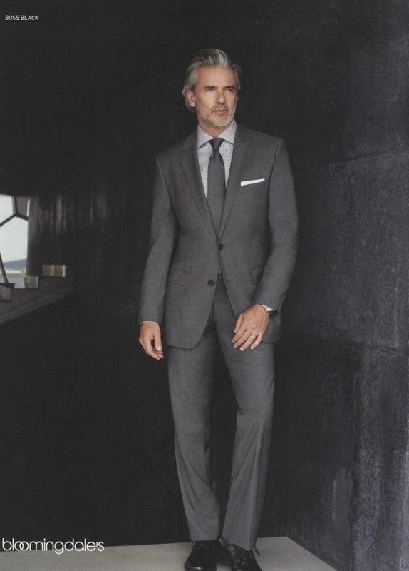 Silver-haired man in a gray suit poses in a dimly lit corridor with gray walls in a full-length studio fashion shot.