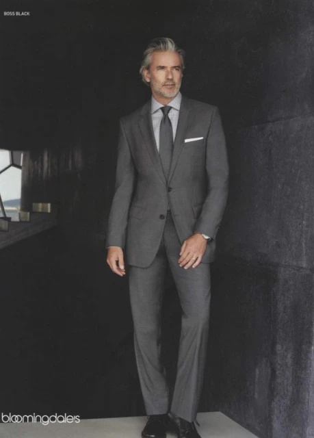 Silver-haired man in a gray suit poses in a dimly lit corridor with gray walls in a full-length studio fashion shot.