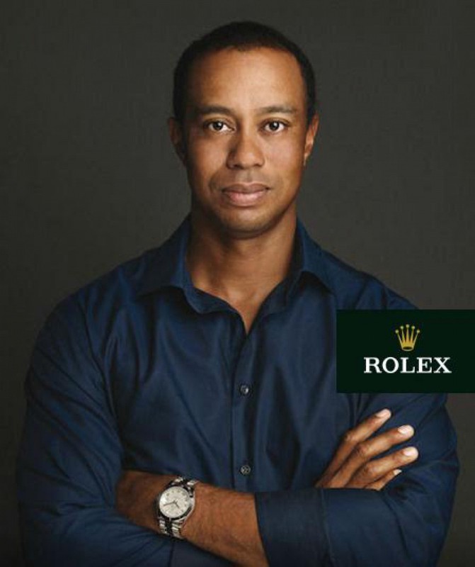 Portrait of Tiger Woods wearing a blue shirt and a Rolex watch, with his arms crossed against a neutral grey backdrop.