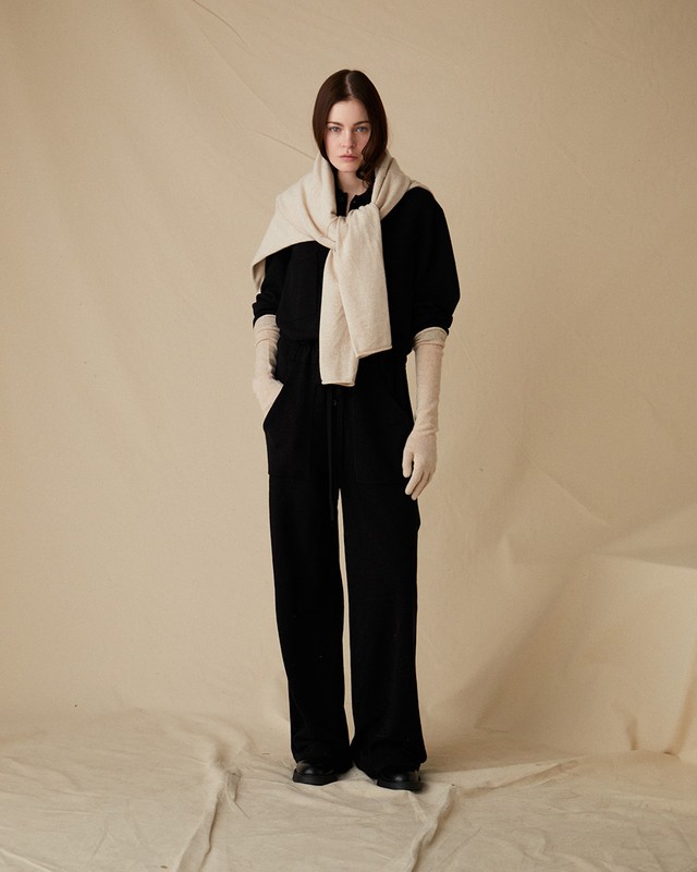 Full shot of a brunette woman wearing a black sweater and pants with beige gloves, and sweater draped over her shoulders in a studio.