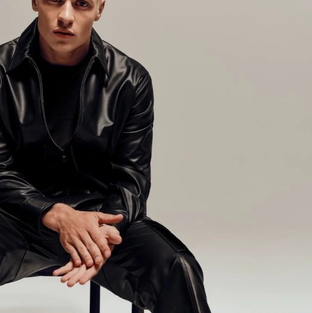 Man posing seated wearing a black leather-like shirt and pants in a studio setting with a neutral background and direct lighting.