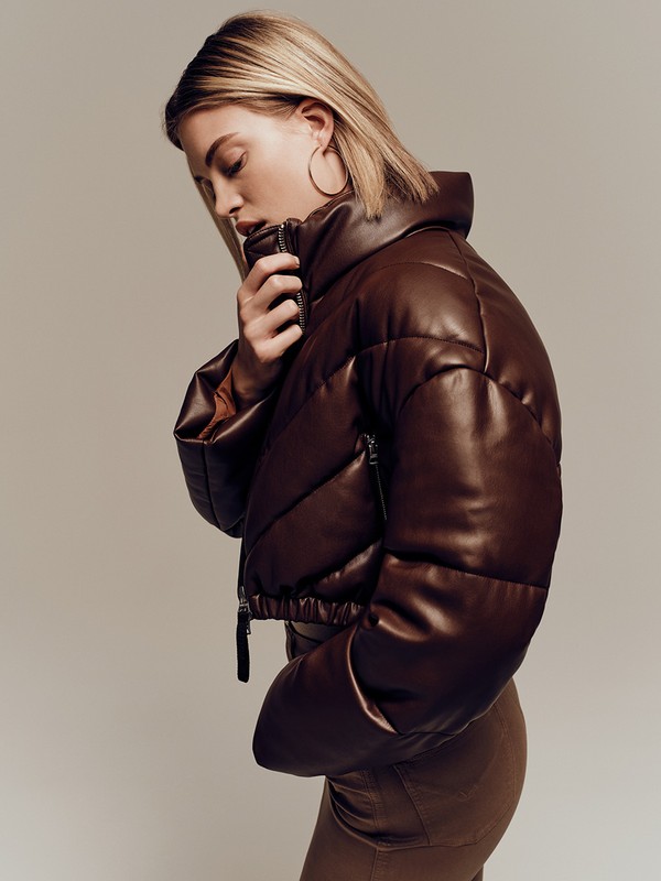 A woman in a stylish brown faux leather puffer jacket poses in a studio with neutral background, showcasing a high fashion editorial look.